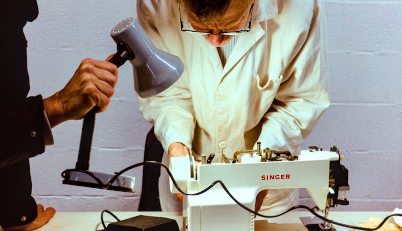 Sur une table une machine à coudre ouverte. Une personne penchée au dessus a les maine dedans et une autre personne éclaire avec une lampe.